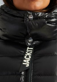 Schwarze, glänzende Pufferjacke mit Kragen, der mit zwei Druckknöpfen mit der Aufschrift "JACKIT" geschlossen ist. Die Jacke hat einen schwarzen Zipper mit einem weißen "JACKIT"-Text.