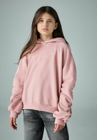 Světle růžová mikina s uvolněným střihem, která má logo na hrudi a žebrované detaily na manžetách. V kombinaci se šedými ošoupanými džíny.