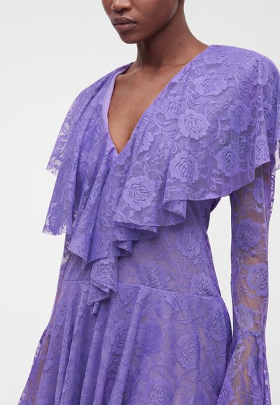 Personne noire portant une robe en dentelle violette avec des motifs floraux, dotée d'un profond décolleté en V et de détails à volants superposés sur la poitrine et les manches.