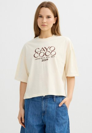 Joven mujer con cabello liso castaño claro lleva una camiseta cremada de gran tamaño con texto "Cayo Coco Jardines del Rey 2009" y vaqueros azules de talle alto.