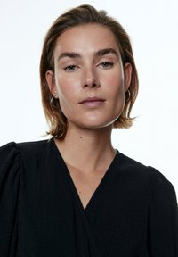 Zwart textuur jurk met een diepe V-hals en pofmouwen, met subtiele stikdetails; model heeft kort haar en ringoorbellen.