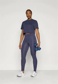 Jūras zila cropped top un augsto jostu legingi, abi izgatavoti no gludas auduma. Tur rokās zilos hanteles, uz kājām baltas sporta kurpes. Neitrāla fonā.