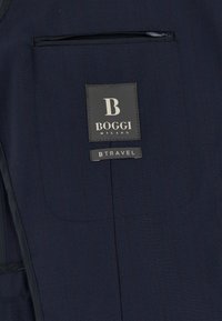 Odevná tkanina námorníckej modrej farby s našitou etiketu "Boggi Milano" a štítkom "B Travel" pri vodorznom vrecku so zipsom.