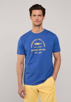 Camiseta de algodón azul con texto y gráficos en amarillo, de manga corta y con cuello redondo. Los acentos incluyen un diseño de tiburón y el nombre de un lugar.