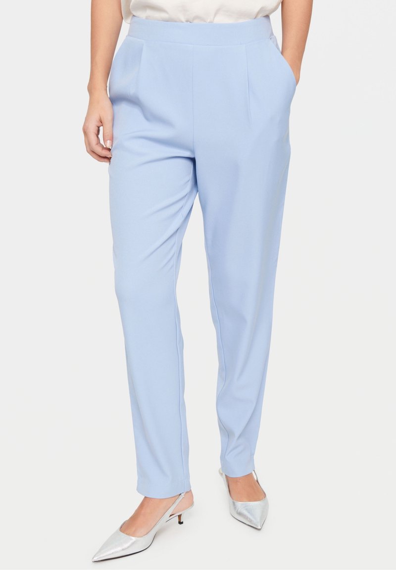 Femme portant un pantalon bleu clair taille haute avec poches latérales et des talons argentés à bride arrière et bout pointu, debout avec une main dans la poche.