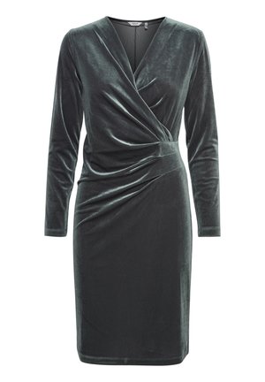 Tamsiai žalia aksominė suknelė su ilgomis rankovėmis, V formos iškirpte ir raukiniais juosmens srityje. Lygi tekstūra ir prigludusi siluetas.