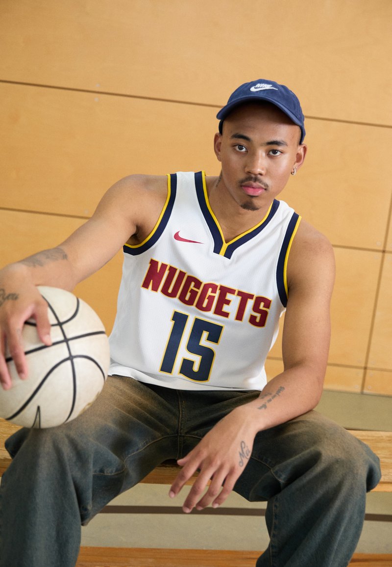 Biela basketbalová dres s nápisom "NUGGETS" a číslom "15" v červenej a modrej farbe, kombinovaný s voľnými džínsami a tmavomodrou čiapkou. Drží basketbal.