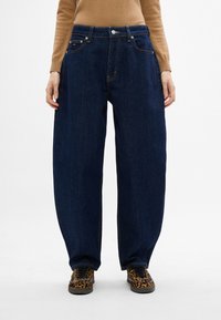 Jeans de mezclilla azul oscuro con un ajuste holgado, cinco bolsillos y costuras doradas. Llevados con zapatillas de deporte con estampado para hacer contraste.