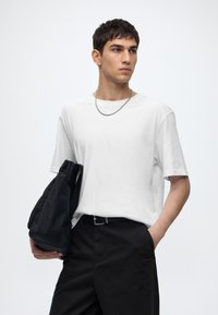 Young man wearing a white t-shirt and black pants holding a black bag with one hand in pocket, looking to the side against a plain background.