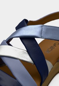 Μπλε και ασημένιο δερμάτινο σανδάλι με σταυρωτούς ιμάντες, με υφασμένο φινίρισμα και καφέ πάτο, σε ανοιχτό φόντο.