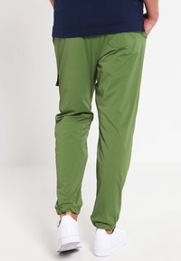 Zelené sportovní kalhoty s elastickými manžetami, bočními kapsami a lehkou, hladkou texturou. Kombinovány s bílými teniskami.