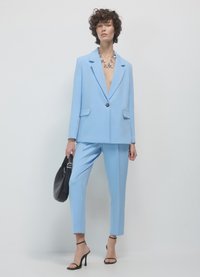 Blazer bleu clair et pantalon assorti en tissu lisse, avec une fermeture à un bouton et deux poches avant. Sac à main noir et talons.