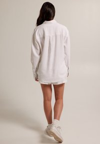 Žena s dlouhými tmavými vlasy, oblečená v oversized bílém tričku, bílých šortkách, bílých ponožkách a bílých teniskách, stojí na jednobarevném pozadí.