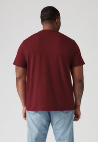 Bourgogne kortærmet t-shirt, lavet af bomuldsmateriale, rund halsudskæring, glat tekstur, vist fra ryggen med blå jeans og synlige tatoveringer.