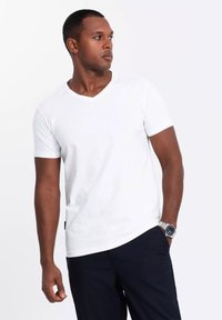 Camiseta blanca de manga corta y escote en V, elaborada con una tela suave. Se usa con pantalones oscuros; presenta una pequeña etiqueta con el logo en el costado. Diseño simple y clásico.