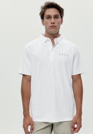 Junger Mann mit heller Haut und braunen Haaren, der ein weißes kurzärmeliges Poloshirt mit kleinem schwarzem Logo auf der Brust trägt und vor einem schlichten Hintergrund steht.