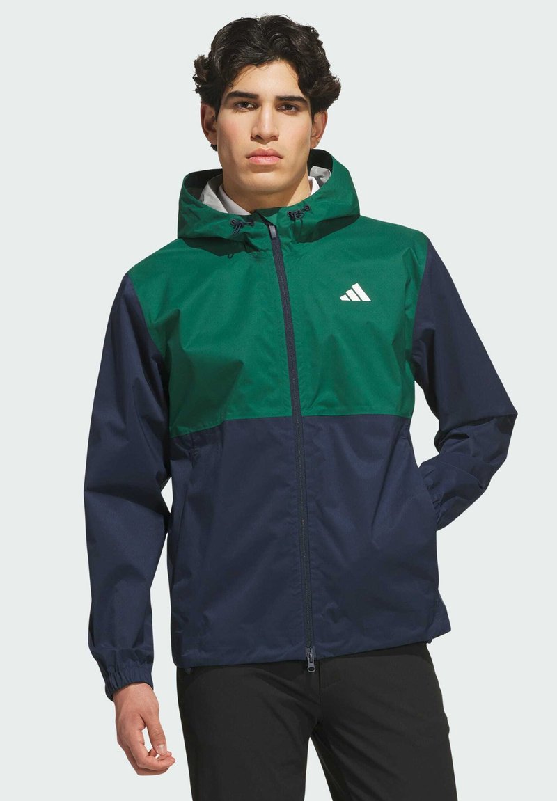 Jeune homme portant une veste à capuche zippée verte et bleu marine avec un logo sportif blanc sur la poitrine, debout devant un fond clair uni.