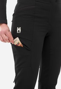 Pantalons noirs d'extérieur avec une finition texturée, dotés d'une poche zippée sur la cuisse et d'une barre de céréales visible partiellement glissée à l'intérieur.