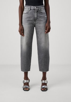 Modelo con vaqueros de lavado gris de cintura alta y corte cropped con ligeros detalles desgastados, combinados con top negro y sandalias con hebilla metálica plateada.