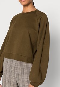 Olivově zelená krátká mikina s raglánovými rukávy, vyrobená z měkké látky. Doplněná kostkovanými kalhotami v neutrálních tónech.