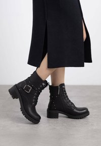 Botas negras de tobillo con un acabado texturizado, diseño con cordones, cremallera lateral y detalle de hebilla, con un tacón grueso y detalles de tachuelas.