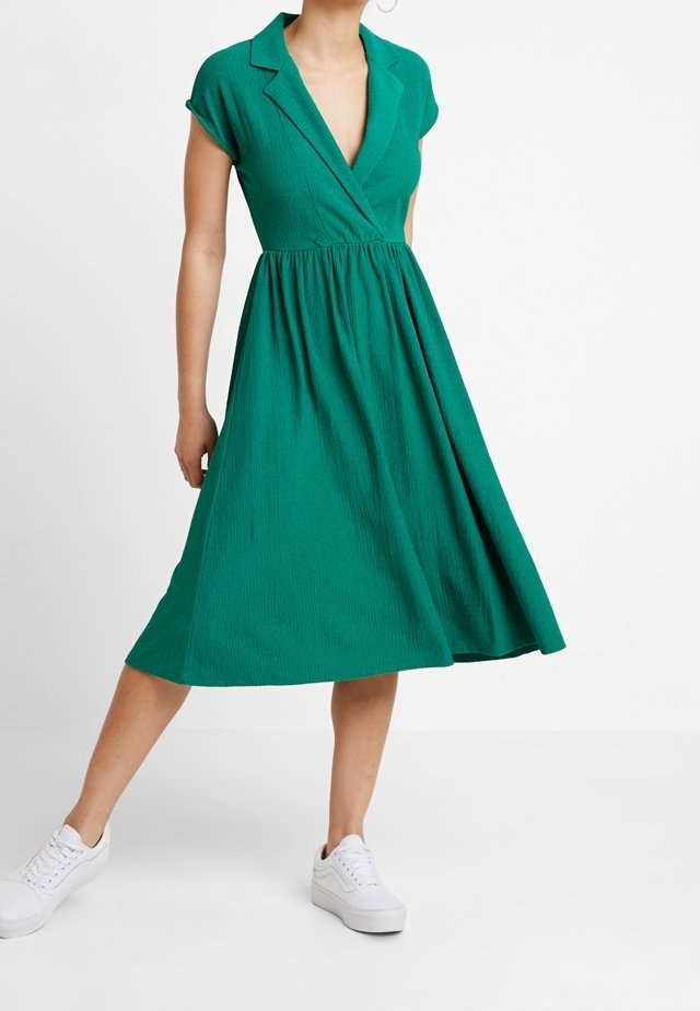 Robe verte à manches courtes, avec un col en V, taille cintrée et jupe évasée. Fabriquée en tissu texturé, avec un design de col et une silhouette simple.