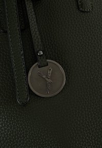 Borsa a mano in pelle verde con finitura texturizzata. Presenta un'etichetta metallica rotonda con un design in legno e un dettaglio della tracolla nera.