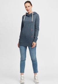 Hettegenser i mørkeblått stoff med lyse snor. Har trekantede brune detaljer og sidelommer. Kombinert med lyseblå jeans.