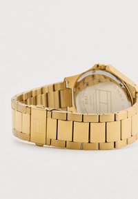 Gold metal link wristwatch band with clasp showing subtle engraved logo, and partially visible round watch case back with engraved text.