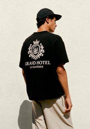 Ung mand iført sort kasket, sort t-shirt med "Grand Hotel Les Sanières" logo på ryggen og lyse beige bukser, stående op ad en hvid væg.