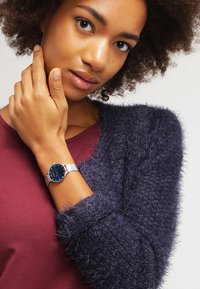 Reloj con correa de malla plateada y esfera de color azul oscuro, que se lleva en una mano contra un suéter texturizado azul oscuro y una parte superior burdeos.
