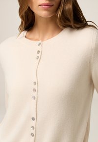 Cárdigan beige hecho de una tela suave, con un cuello redondo y seis botones decorativos de diversos colores en la parte delantera.