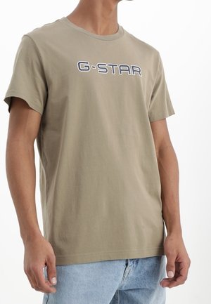 Man wearing beige G-STAR logo t-shirt and light blue jeans, standing with relaxed posture against white background.