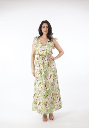 Vrouw staat met hand op heup, draagt een mouwloze, bloemenprint groene en roze maxi-jurk en sandalen met doorzichtige bandjes tegen een witte achtergrond.