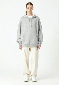 Felpa grigia con cappuccio e coulisse, tasca frontale e polsini a costine, abbinata a pantaloni cargo bianchi con tasche laterali e scarpe da ginnastica bianche.