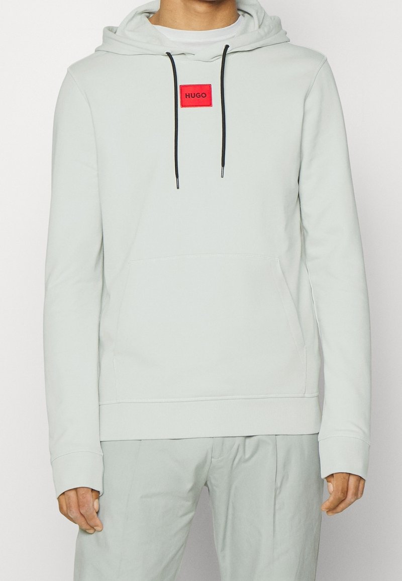 Sweat à capuche gris clair avec cordons noirs et un patch logo rectangulaire rouge portant l'inscription « HUGO » sur la poitrine, porté avec un pantalon gris assorti.