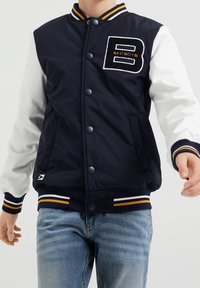 Person som bär en marinblå och vit varsity-jacka med randiga ärmslut och en "B Bad Boys"-patch på bröstet, i kombination med ljusblå jeans.