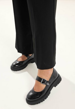 Füße in schwarzen Mary-Jane-Schuhen mit klobigen Sohlen und silbernen Schnallenriemen, kombiniert mit schwarzen geraden Hosen.