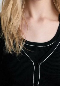 Cardigan noir à côtes avec bord blanc, présentant un col en V et une fermeture à boutons. En dessous, un haut noir à col arrondi est visible.