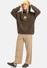 Brauner Kapuzenpullover mit einer Vorderasche und einem orangenen Logo, kombiniert mit beigen und weißen karierten weiten Hosen und braunen Schuhen.