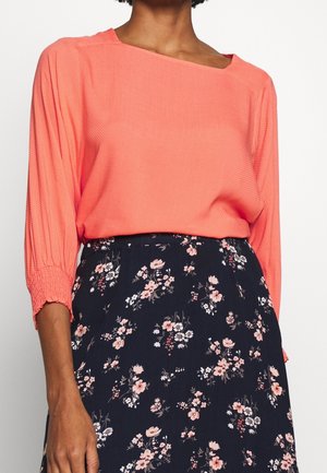 Vrouw draagt een koraalkleurige blouse met lange mouwen en geplooide manchetten en een zwarte rok met roze en witte bloemenprint.