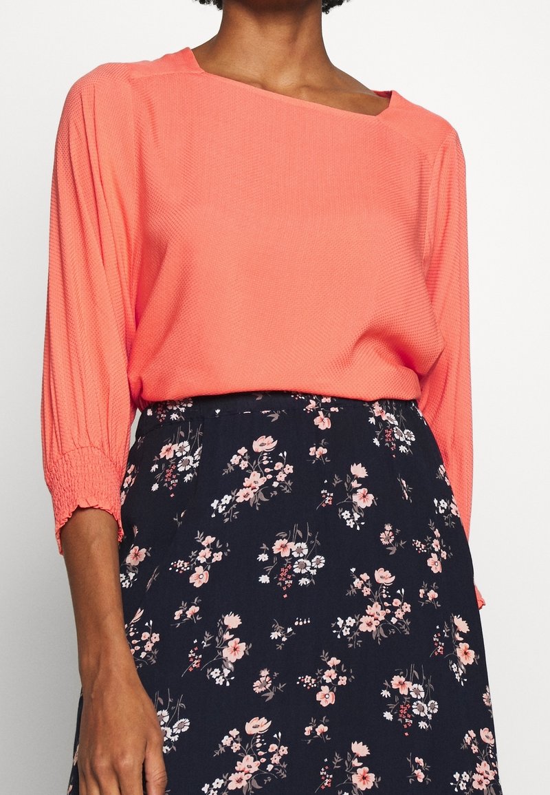 Femme portant un chemisier corail à manches longues avec des poignets froncés et une jupe noire à motif floral rose et blanc.