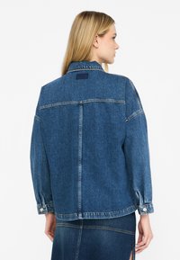 Giubbotto di denim blu scuro con vestibilità rilassata, caratterizzato da un ampio giromanica sul retro e polsini con bottoni. Presenta dettagli di cucitura sottili.