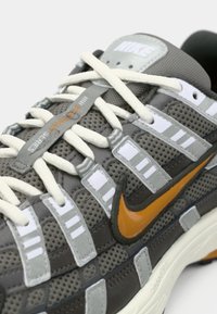 Nike αθλητικό παπούτσι με γκρι διχτυωτό επάνω μέρος και λευκές και γκρι λεπτομέρειες, πορτοκαλί λογότυπο swoosh και υφασμάτινα κορδόνια με υφή. Σημαντική επένδυση στην περιοχή του αστραγάλου.