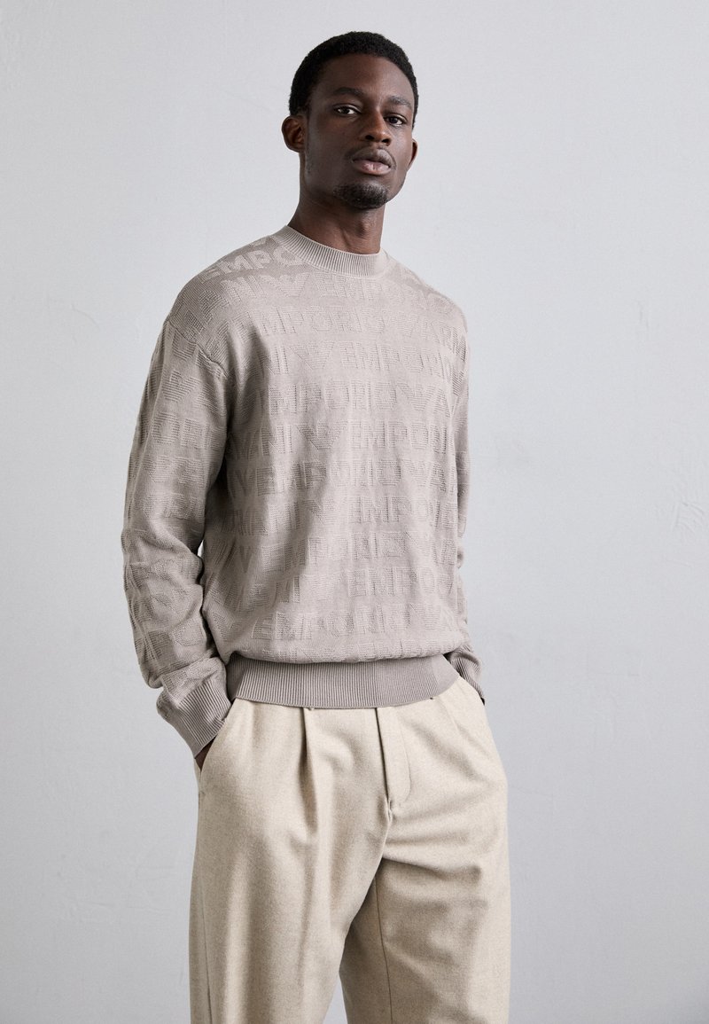 Pullover beige chiaro lavorato a maglia con dettaglio del logo testurizzato, colletto rotondo e polsini a costine, abbinato a pantaloni beige a pieghe.