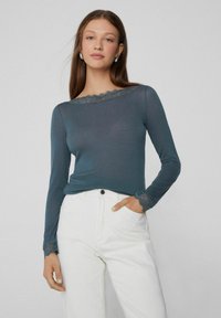 Junge Frau in einem türkisfarbenen Langarmoberteil mit Spitzenbesatz am Ausschnitt und an den Säumen, weißen hoch taillierten Hosen, die mit einer Hand in der Tasche steht.