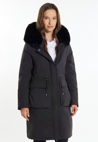 Cappotto isolato nero con colletto in pelliccia artificiale, chiusure con zip e bottoni a pressione, due tasche frontali, vita regolabile con cordino e lunghezza fino al ginocchio.