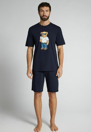 Mann trägt ein marineblaues T-Shirt mit einem Teddybär, der ein Eis hält, und passende marineblaue Shorts, steht barfuß vor einem schlichten Hintergrund.