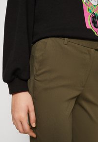 Olivgröna byxor med en slät yta och sidofickor, kombinerat med en svart sweatshirt med en färgglad grafisk design.