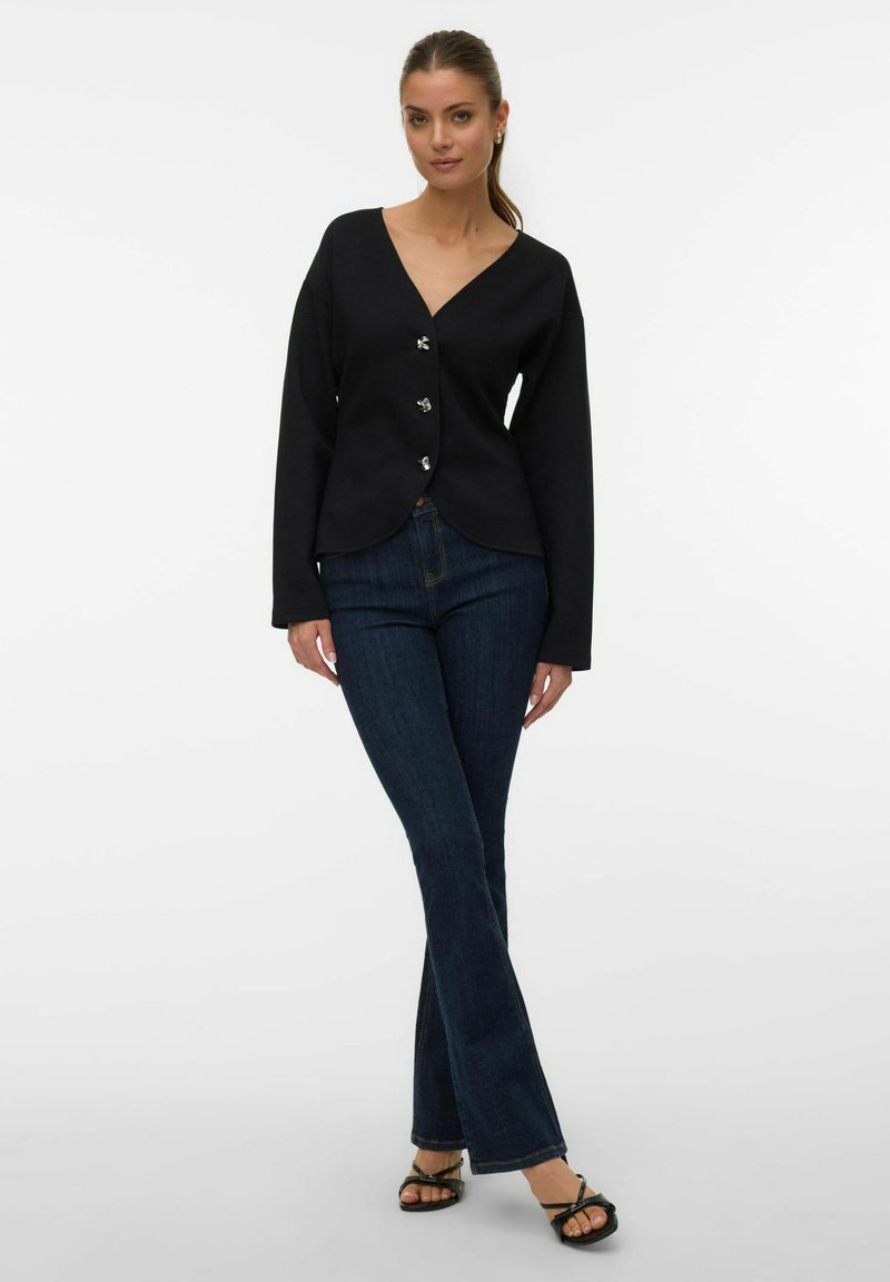 Femme portant un chemisier noir à col en V avec des boutons, un jean bootcut bleu foncé et des sandales à talons ouvertes noires, debout sur un fond blanc.